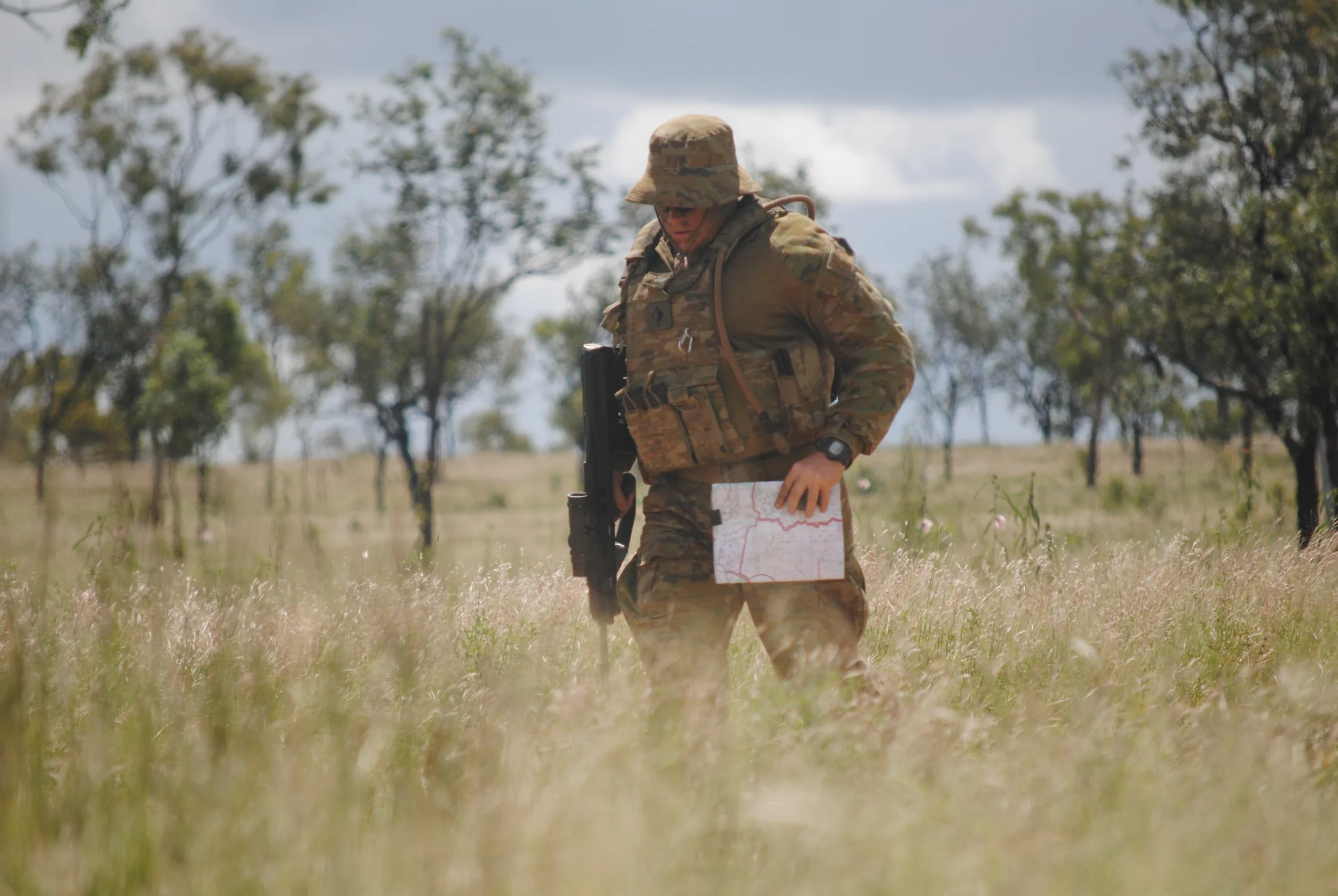 A single soldier in field uniform standing in dry grassland, reading a map during a navigation exercise.