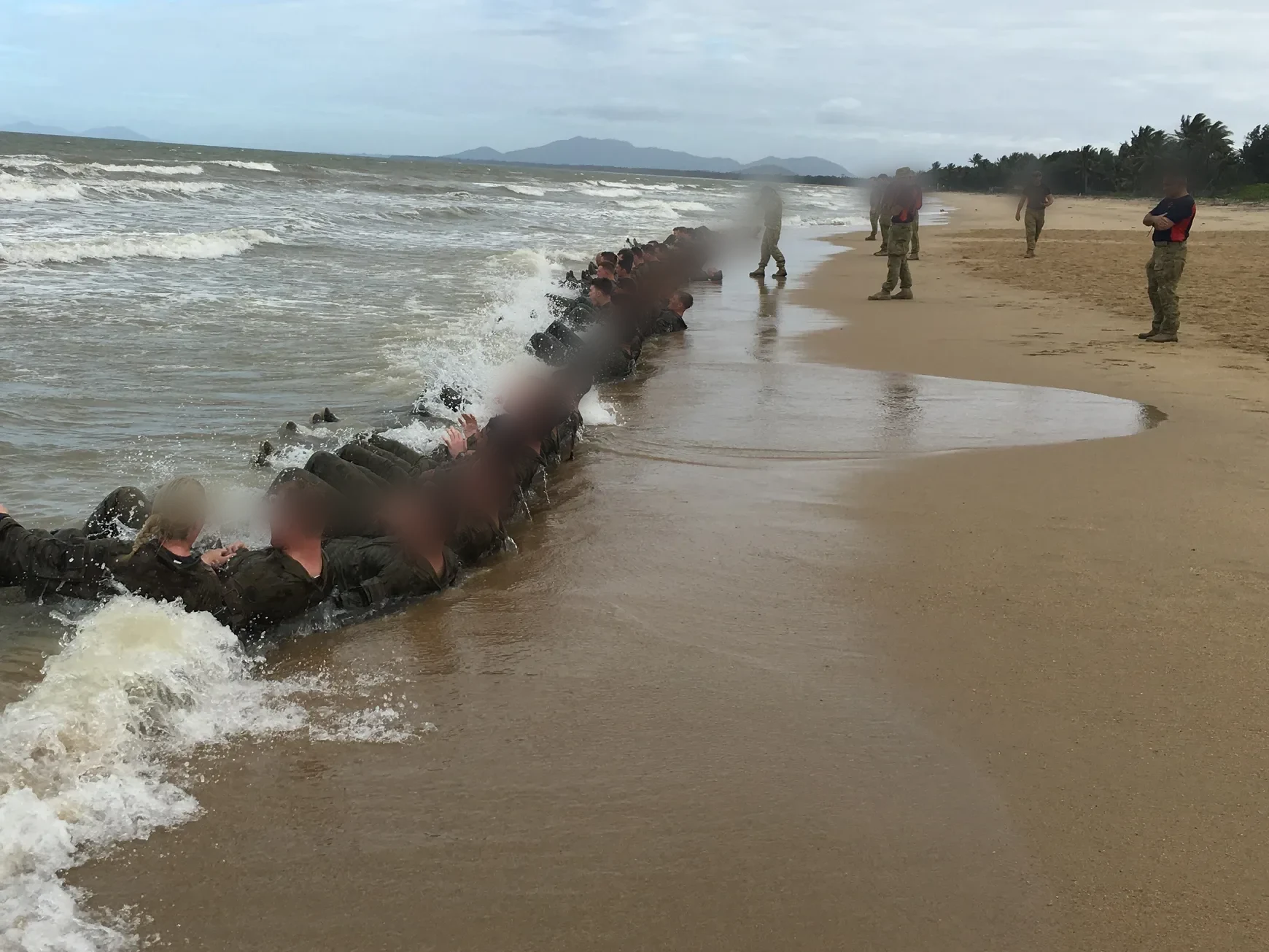A long line of soldiers linked together doing group physical training in the shore break, an instructor standing over them, faces obscured.