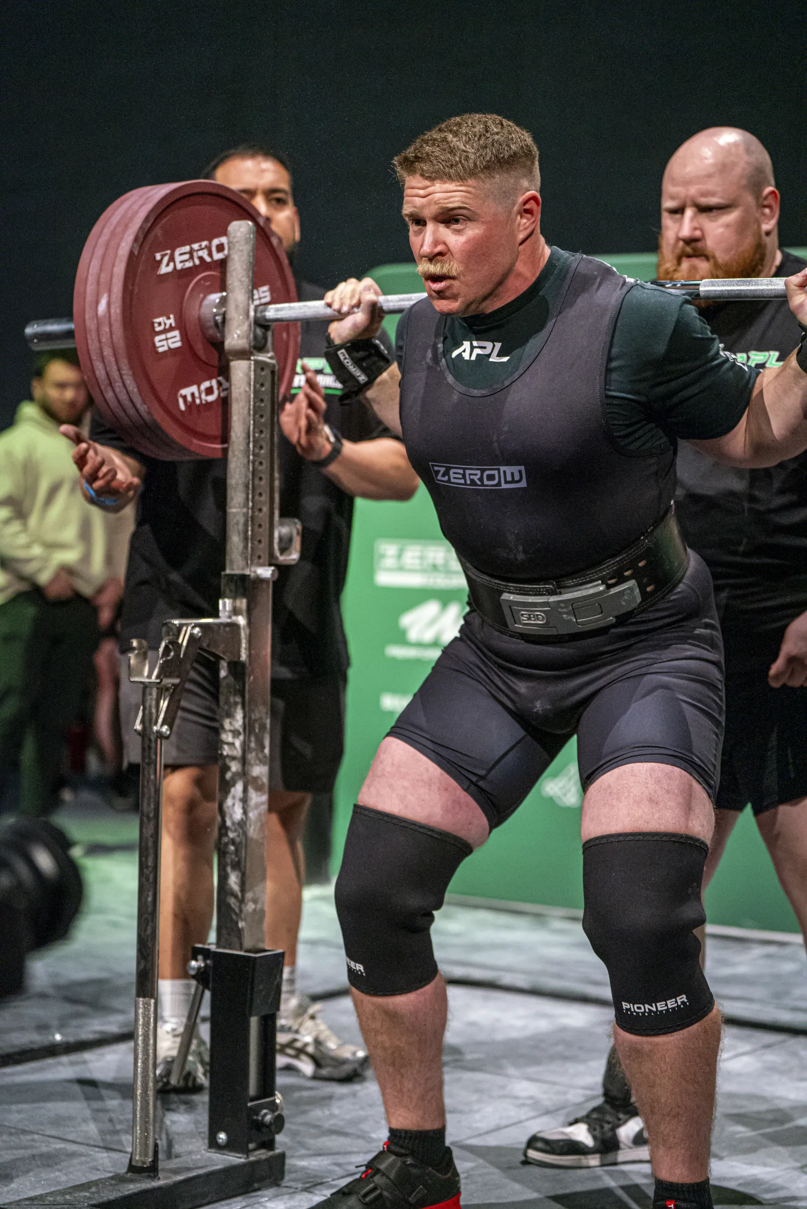 A lifter walking a loaded bar out of the rack at a competition, spotters behind.