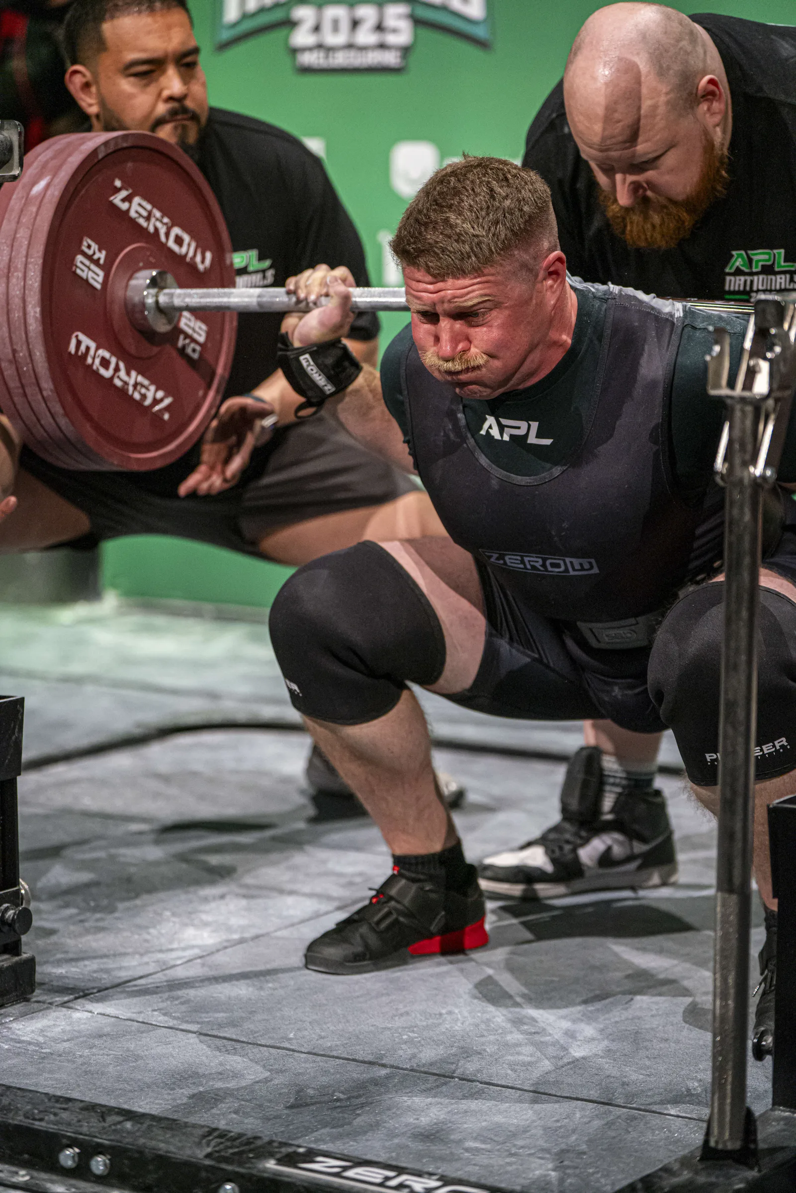 The coach at the bottom of a competition squat, bar loaded with red plates, on a lit platform.