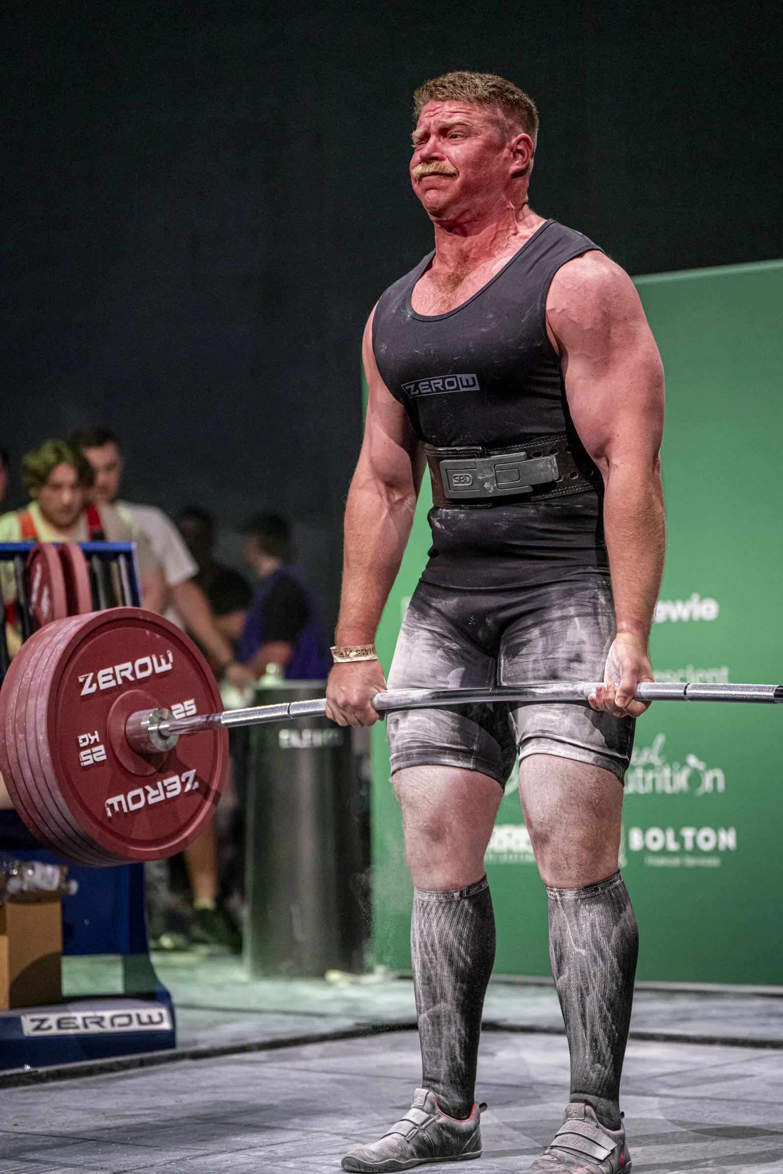 Abe Dearmer at lockout on a competition deadlift, chalk on his hands and singlet, on a powerlifting platform.