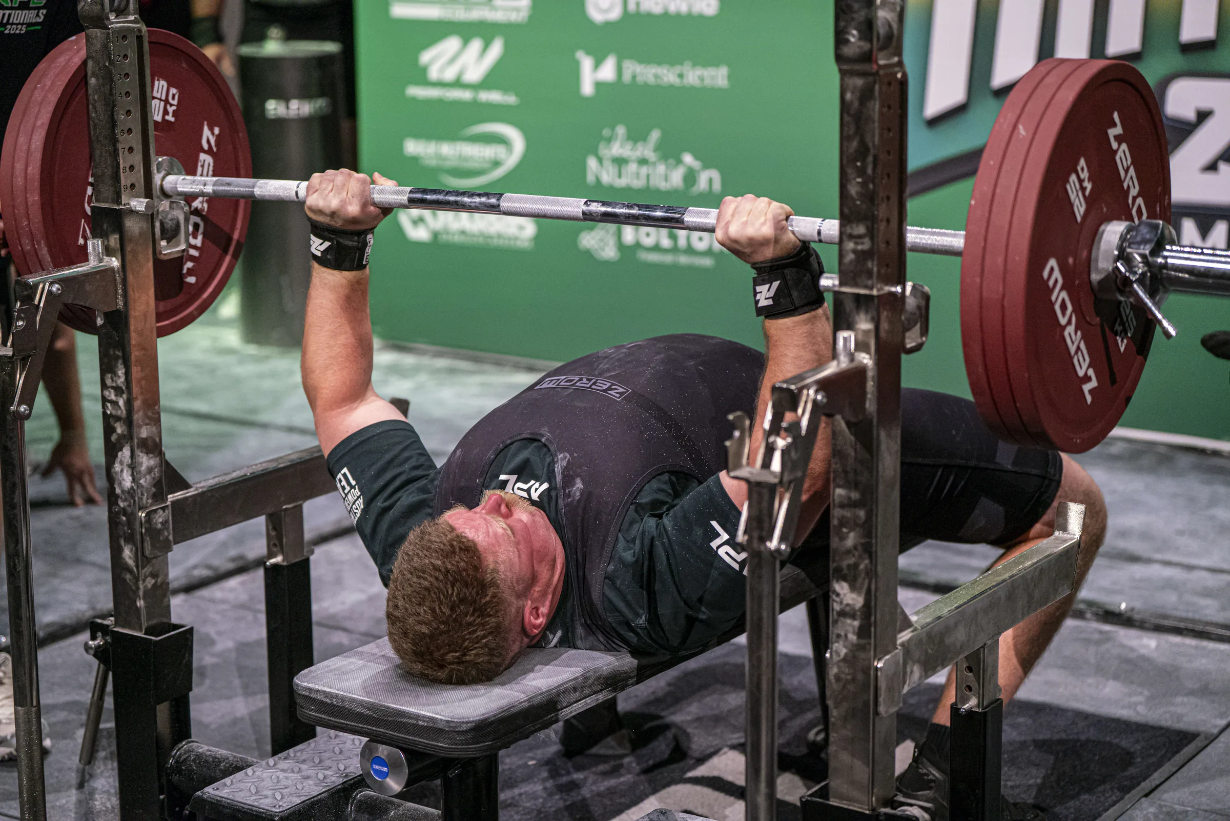 A competition bench press at lockout, spotters either side of the loaded bar.